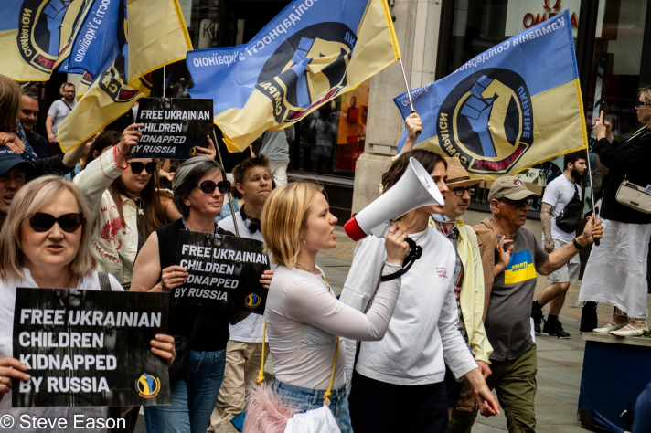 A group of protestors holding signs that read 'FREE UKRAINIAN CHILDREN KIDNAPPED BY RUSSIA' and waving flags, with one person using a megaphone to speak.